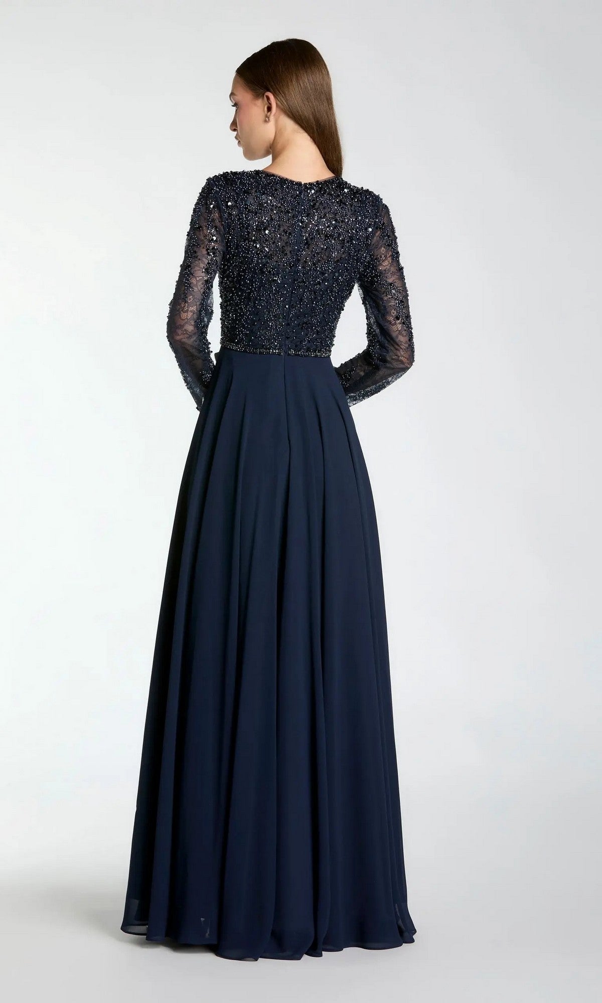A woman is pictured from behind, wearing the Mac Duggal 21316 long formal dress. The floor-length navy gown features an embellished bodice and sheer long sleeves, paired with a flowing, pleated chiffon A-line skirt that enhances the fitted top. The background is plain and light-colored.